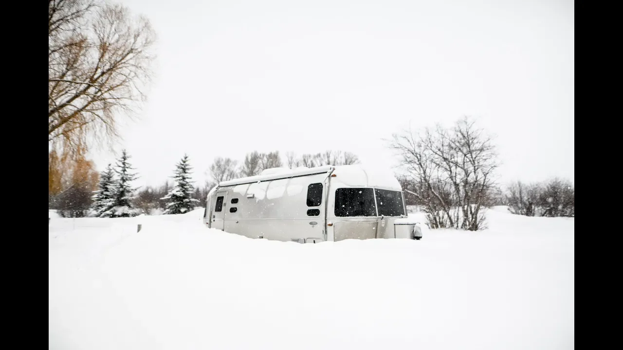 Silver camper trailer partially buried in deep snow, surrounded by snow-covered trees and a bleak winter landscape.