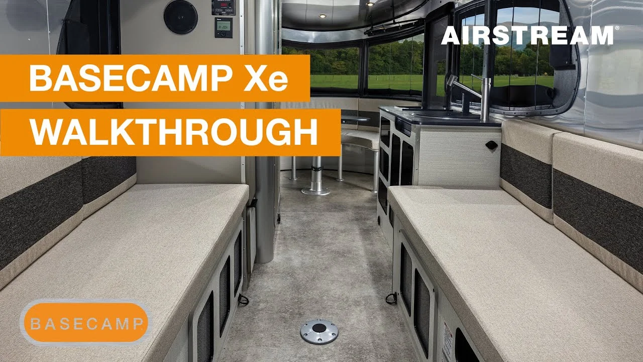 Interior view of an Airstream Basecamp Xe camper, featuring seating, large windows, and compact kitchen. Text overlay: "Basecamp Xe Walkthrough".