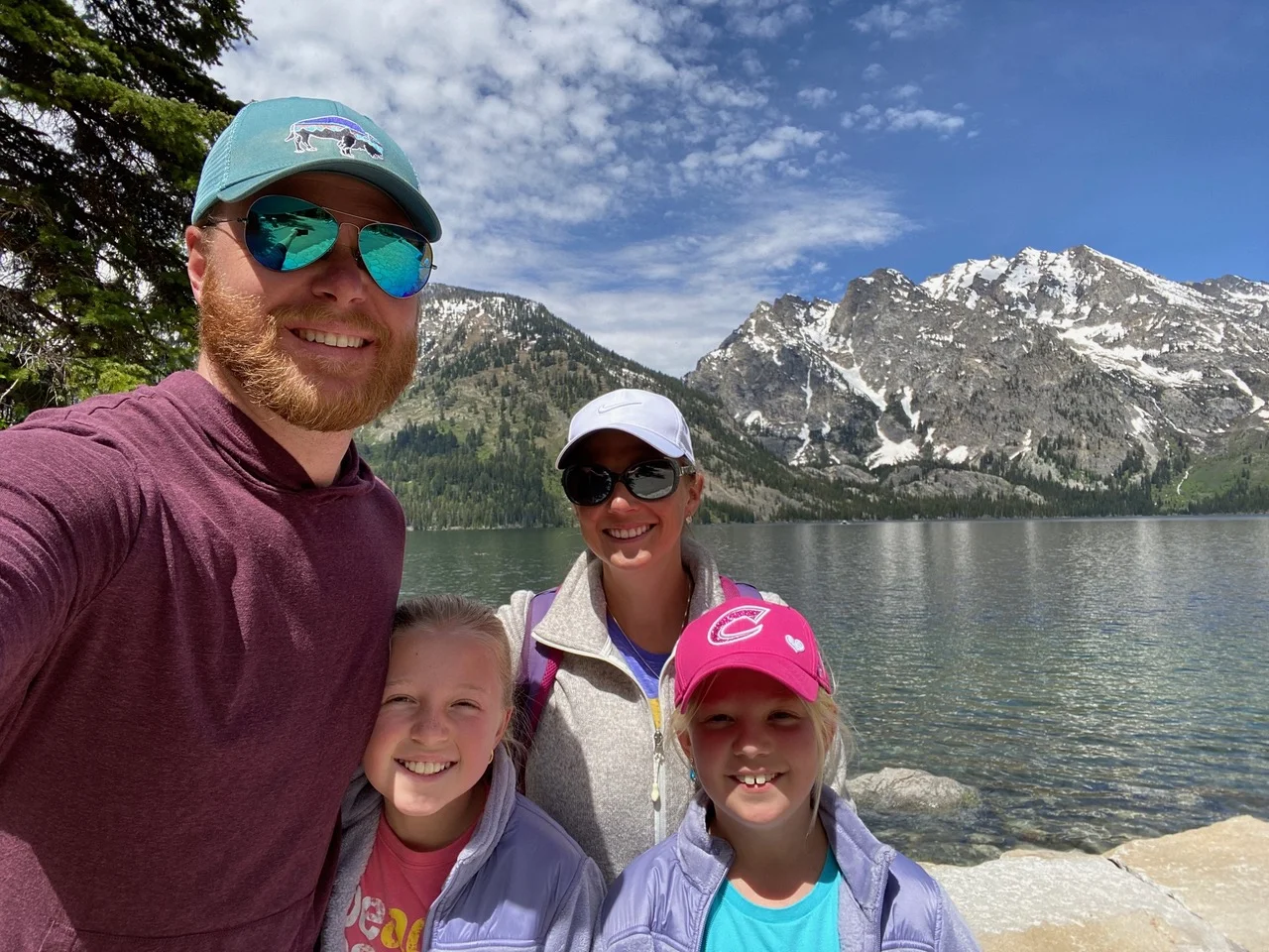 A family of four posing by a mountain lake, with snow-capped peaks in the background. The sky is partly cloudy.