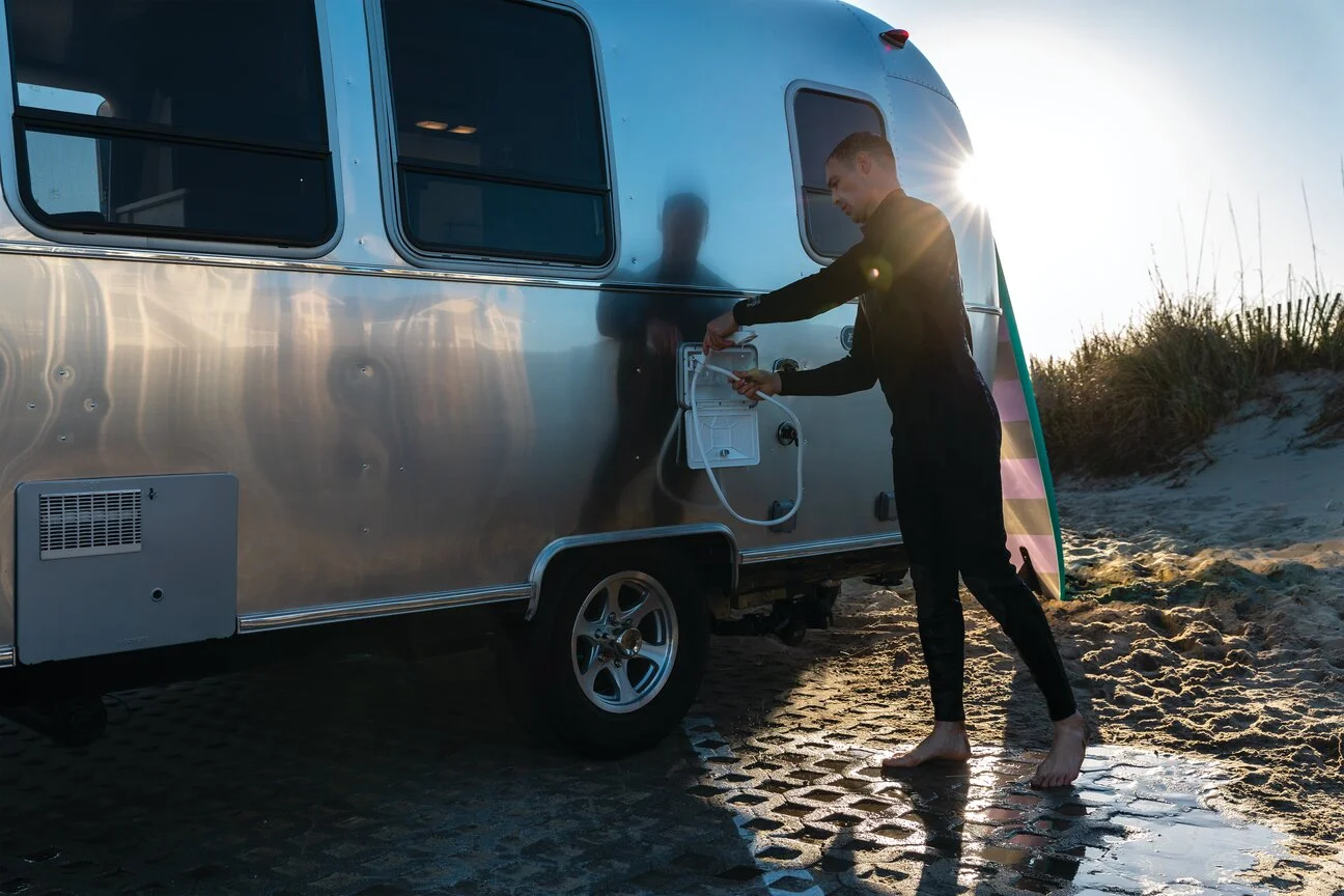 A man in a wetsuit rinses off with a shower attached to a silver trailer, with sunlight and sand dunes in the background.