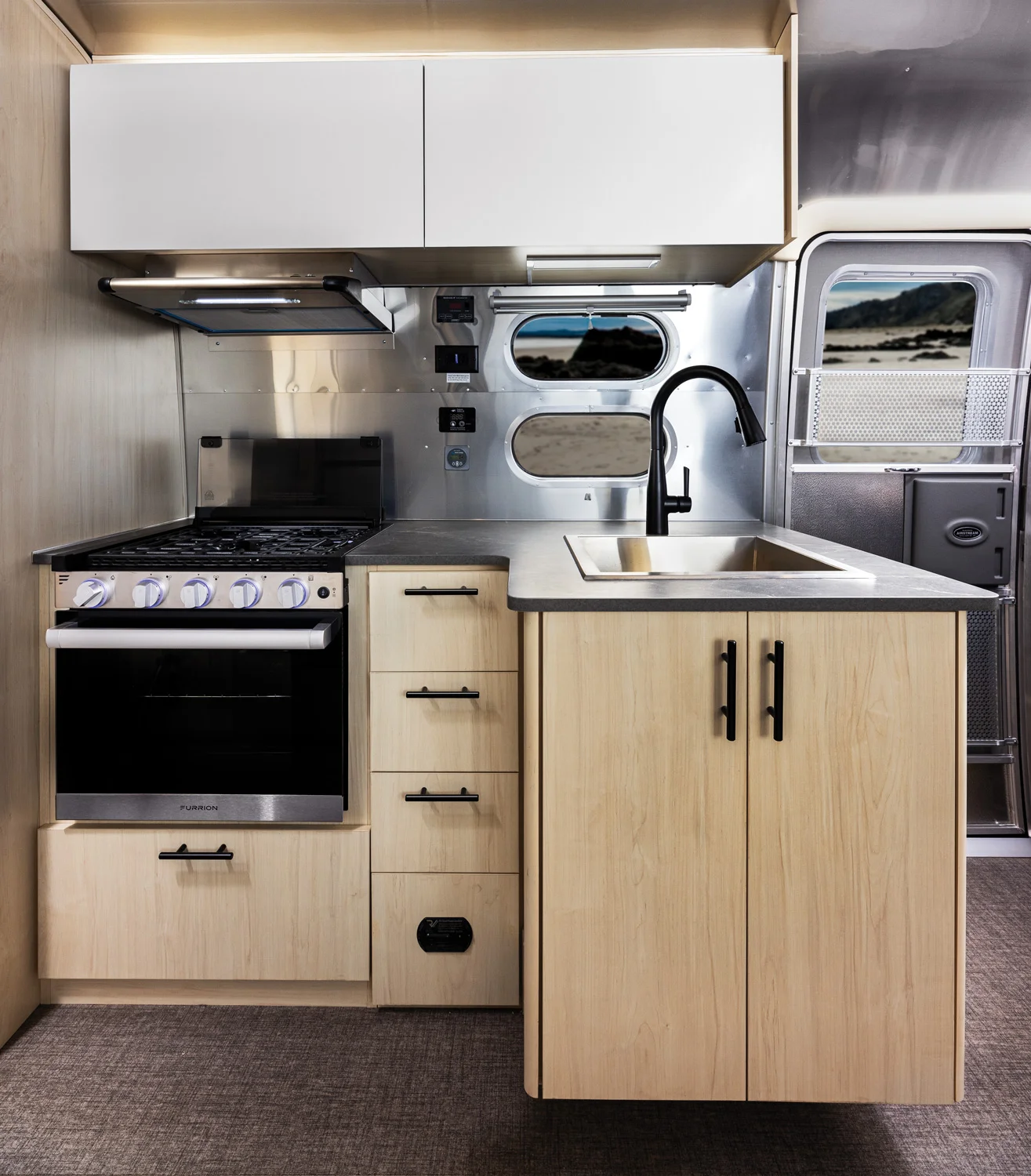 Modern RV kitchen with light wood cabinets, stainless steel sink, black faucet, stove, and built-in oven. Metal wall and window visible.
