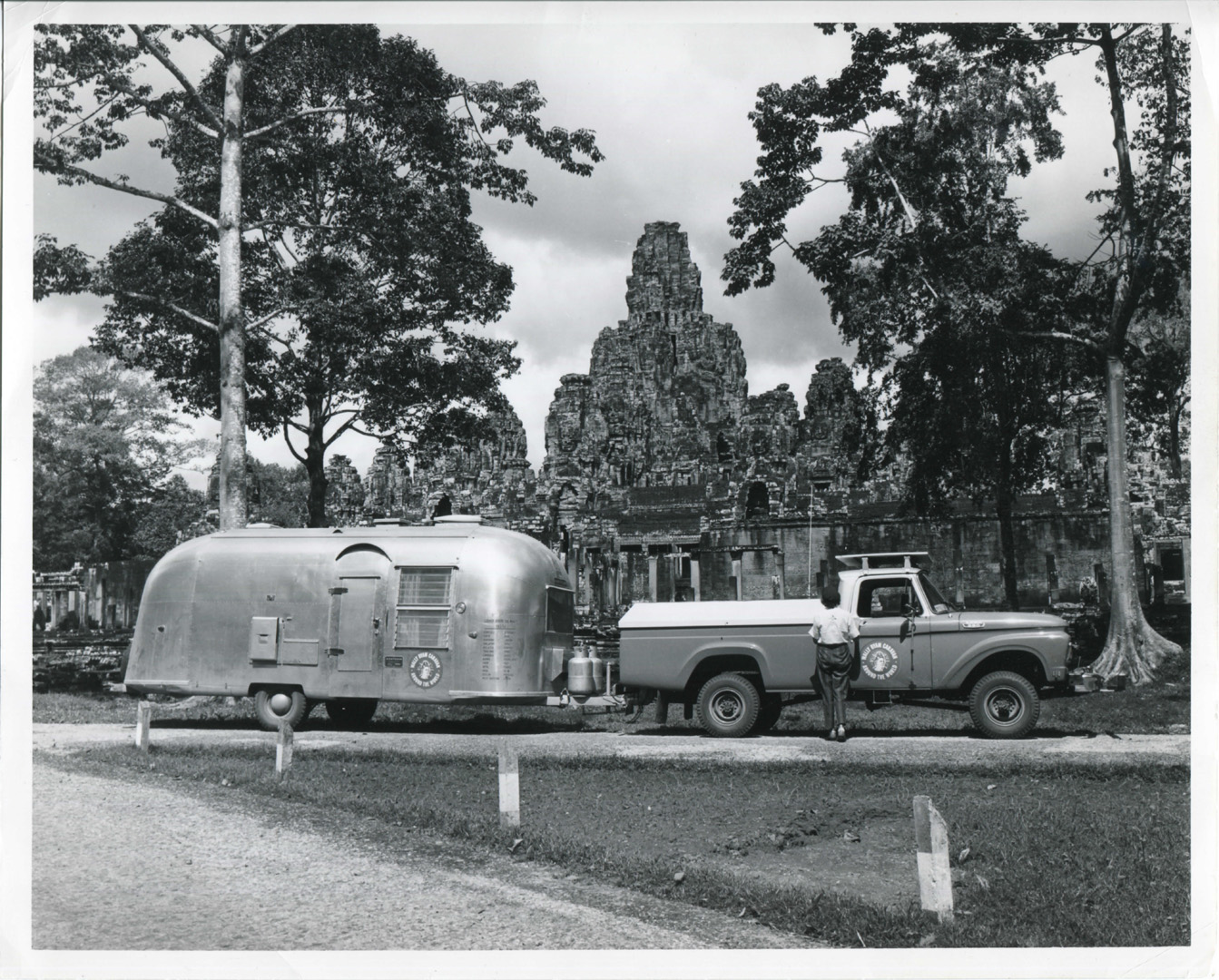 A black-and-white photo of a vintage truck towing an Airstream trailer parked in front of ancient temple ruins surrounded by tall trees. A person stands beside the truck, and dramatic clouds fill the sky.