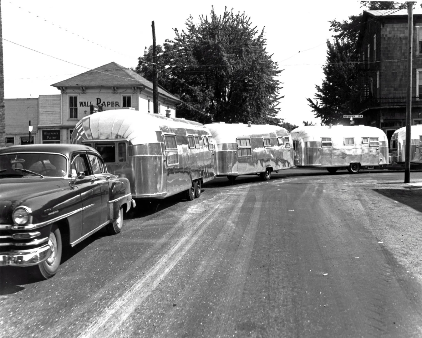 Vintage car towing shiny aluminum trailers on a quiet street with a "Wallpaper & Paint" store in the background.