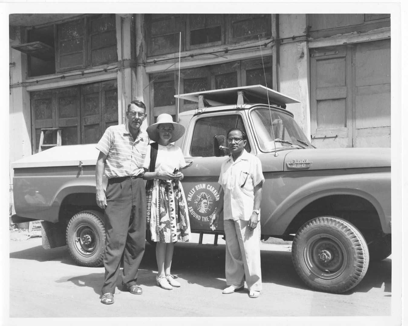 Three people stand in front of a vintage Ford pickup truck with a logo on the door, parked near a building with large doors. The woman in the center wears a hat, and the men wear short-sleeve shirts and glasses.