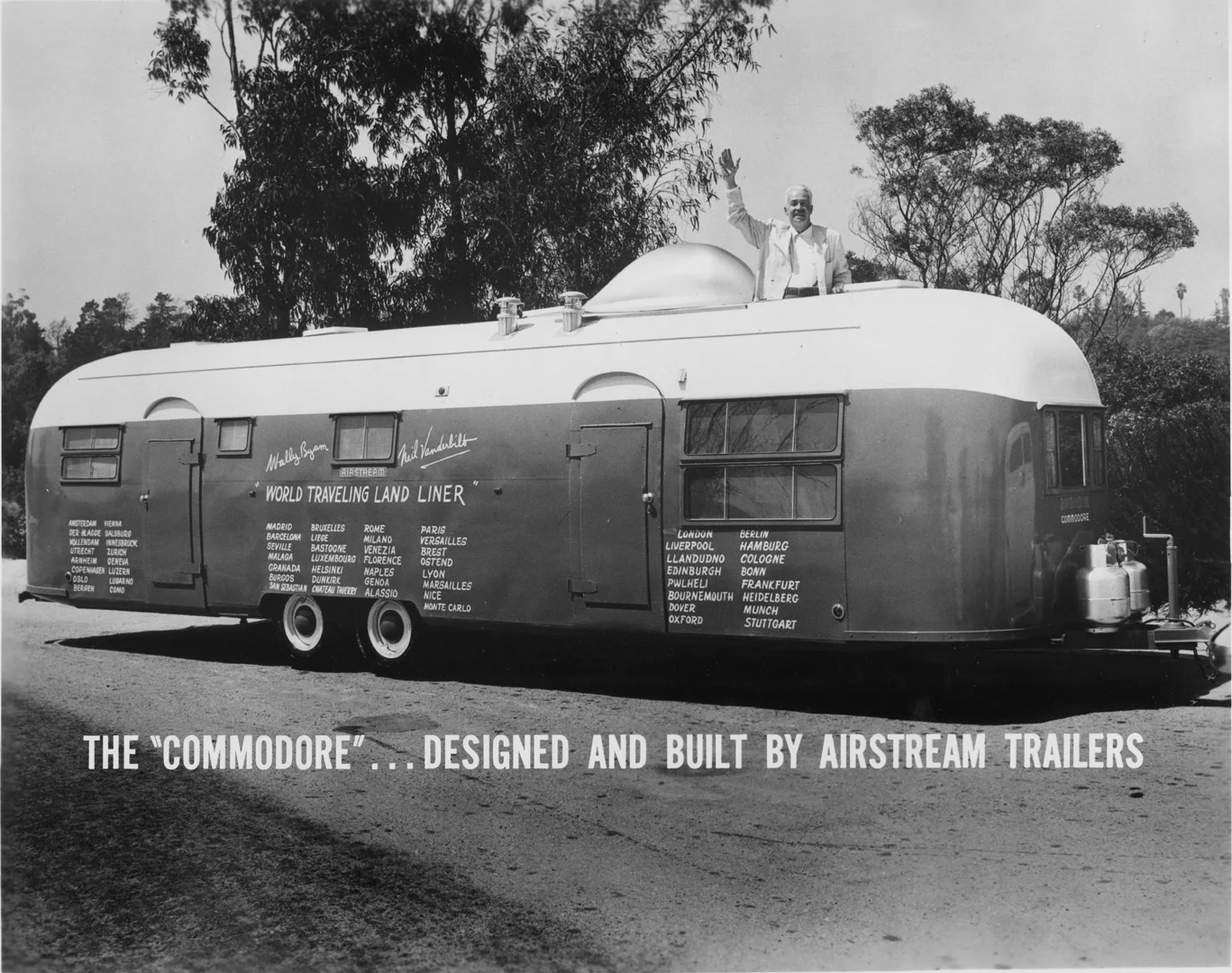 Vintage Airstream trailer labeled "Commodore" with travel destinations listed, man standing on top waving, trees in background.