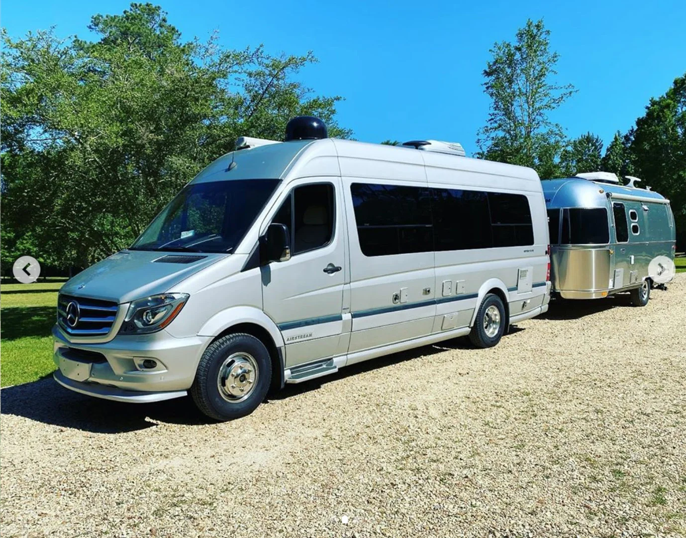 Silver camper van towing a small silver Airstream trailer on a gravel path with trees and blue sky in the background.