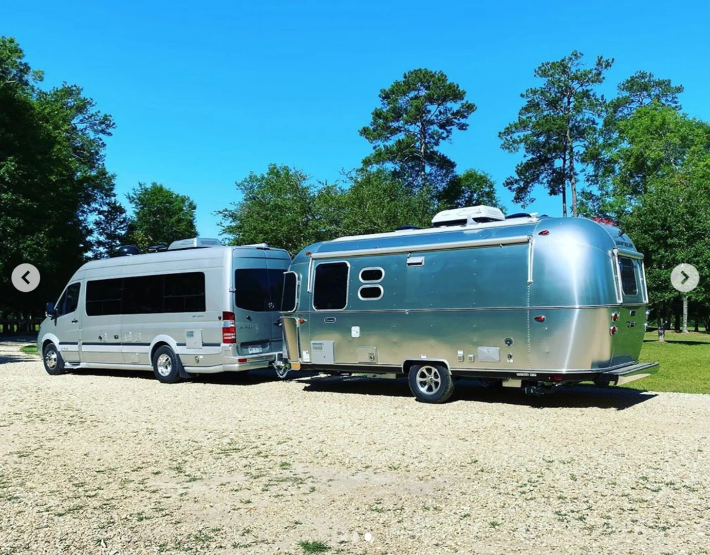 Silver camper van towing a shiny Airstream trailer on a gravel road, with trees and a clear blue sky in the background.