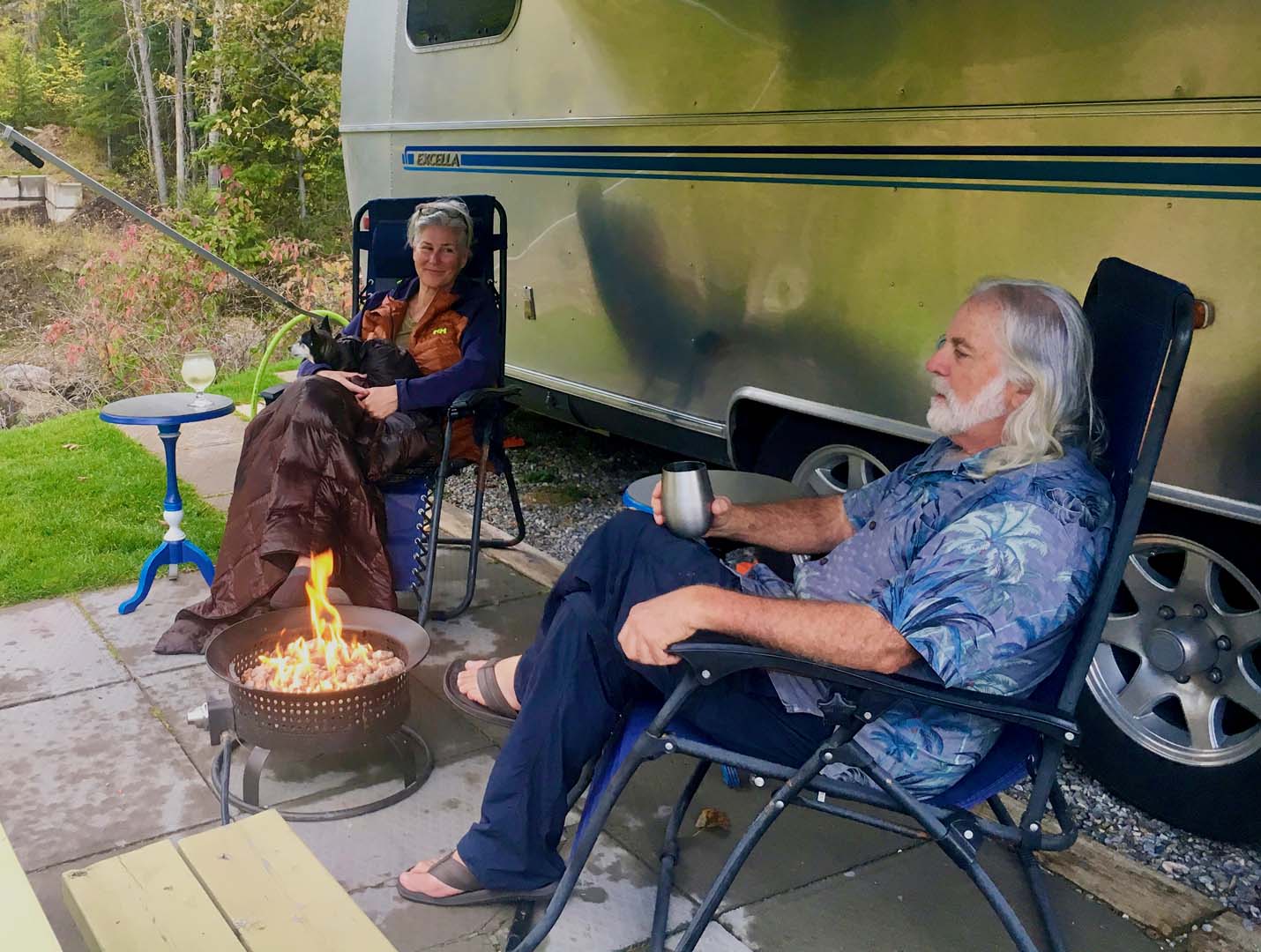 A man and woman relax in chairs by a small fire pit, in front of a trailer, surrounded by greenery.