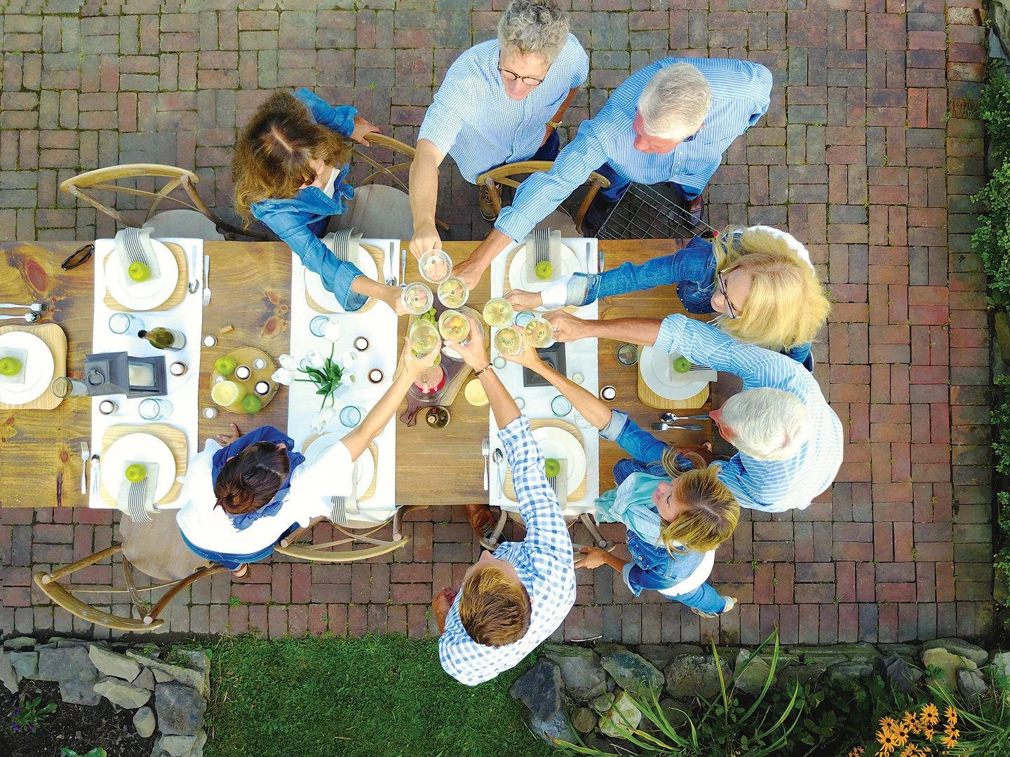A group of people gathered around an outdoor table raise their glasses in a toast. The table is set for a meal, viewed from above, on a brick patio with greenery on the side.