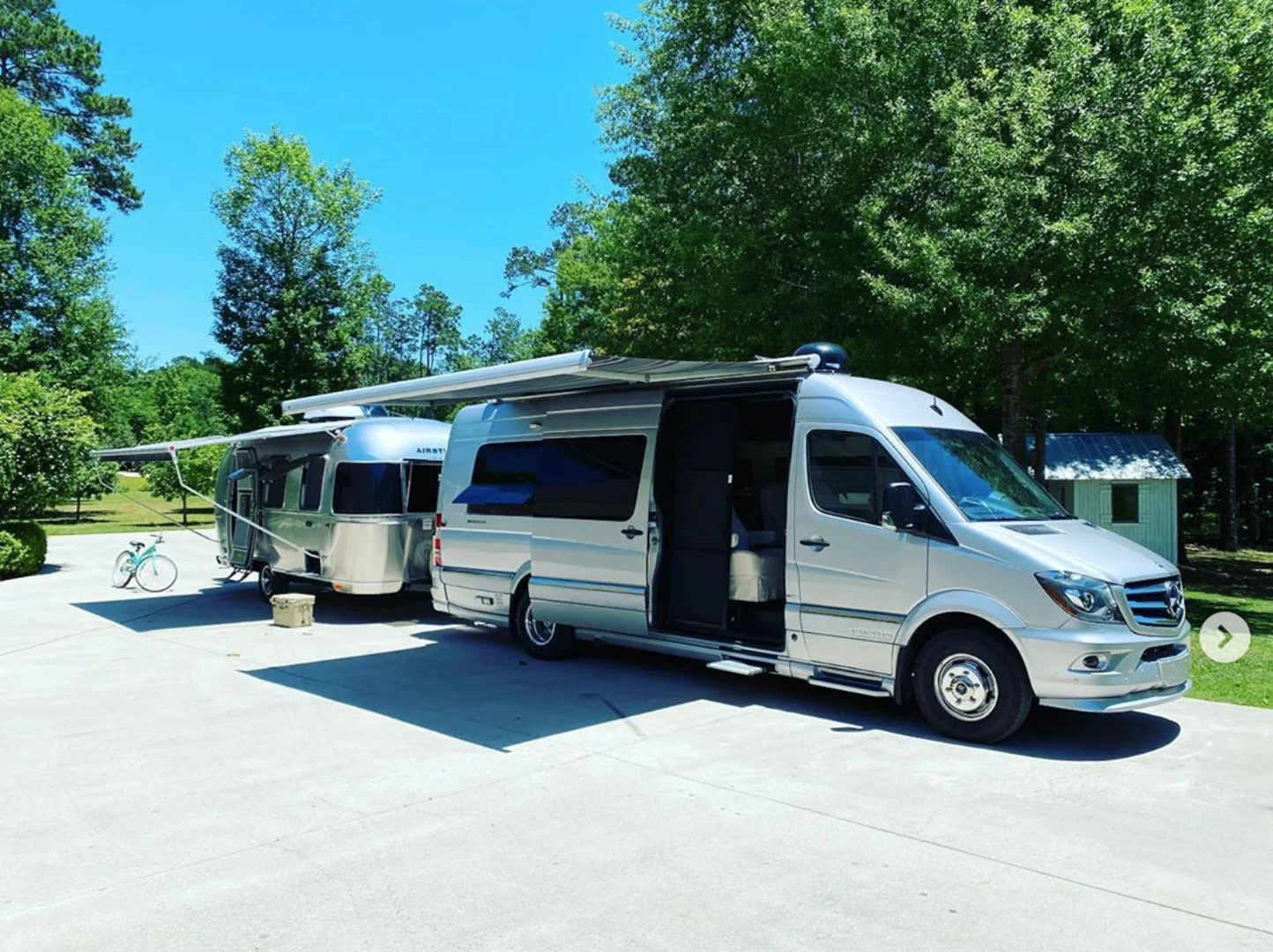 A silver camper van with an extended awning is parked on a driveway, towing a shiny silver trailer. A blue bicycle is nearby under a clear blue sky.