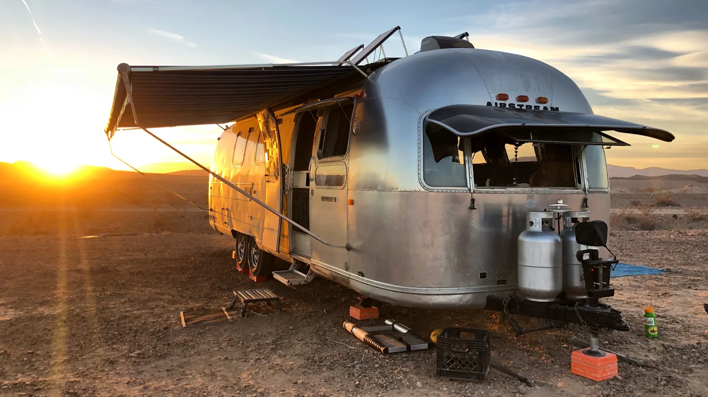 A shiny Airstream trailer with its awning extended is parked on rocky ground at sunset, surrounded by desert landscape and mountains in the distance.