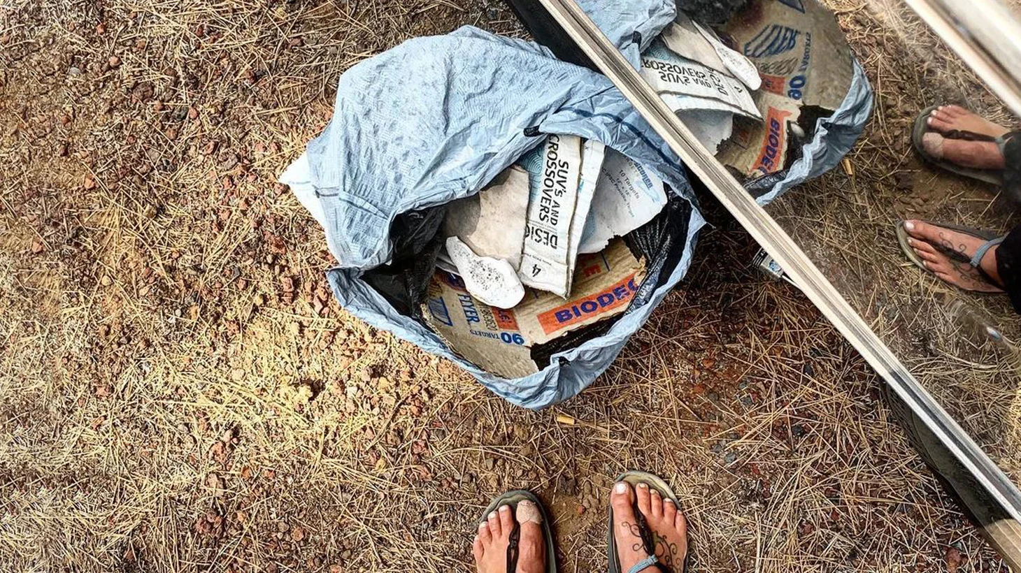 A person wearing sandals stands near a bag filled with various items, including a piece labeled BIODEG, on dry, patchy ground. The scene is partially reflected in a shiny, metallic surface.