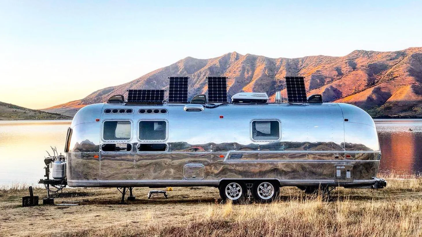 A shiny silver travel trailer with solar panels on its roof is parked by a calm lake, with mountains in the background and dry grass in the foreground at sunset.