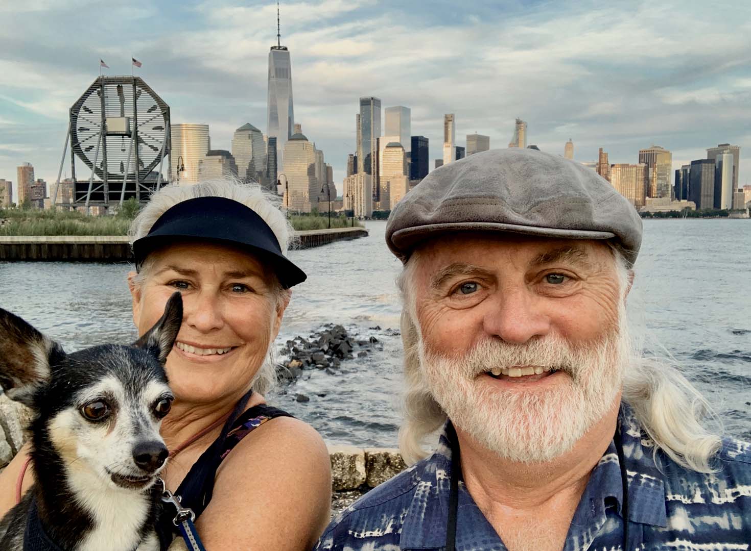 A smiling couple with a small dog takes a selfie in front of a city skyline across the water, with a ferris wheel and skyscrapers visible.