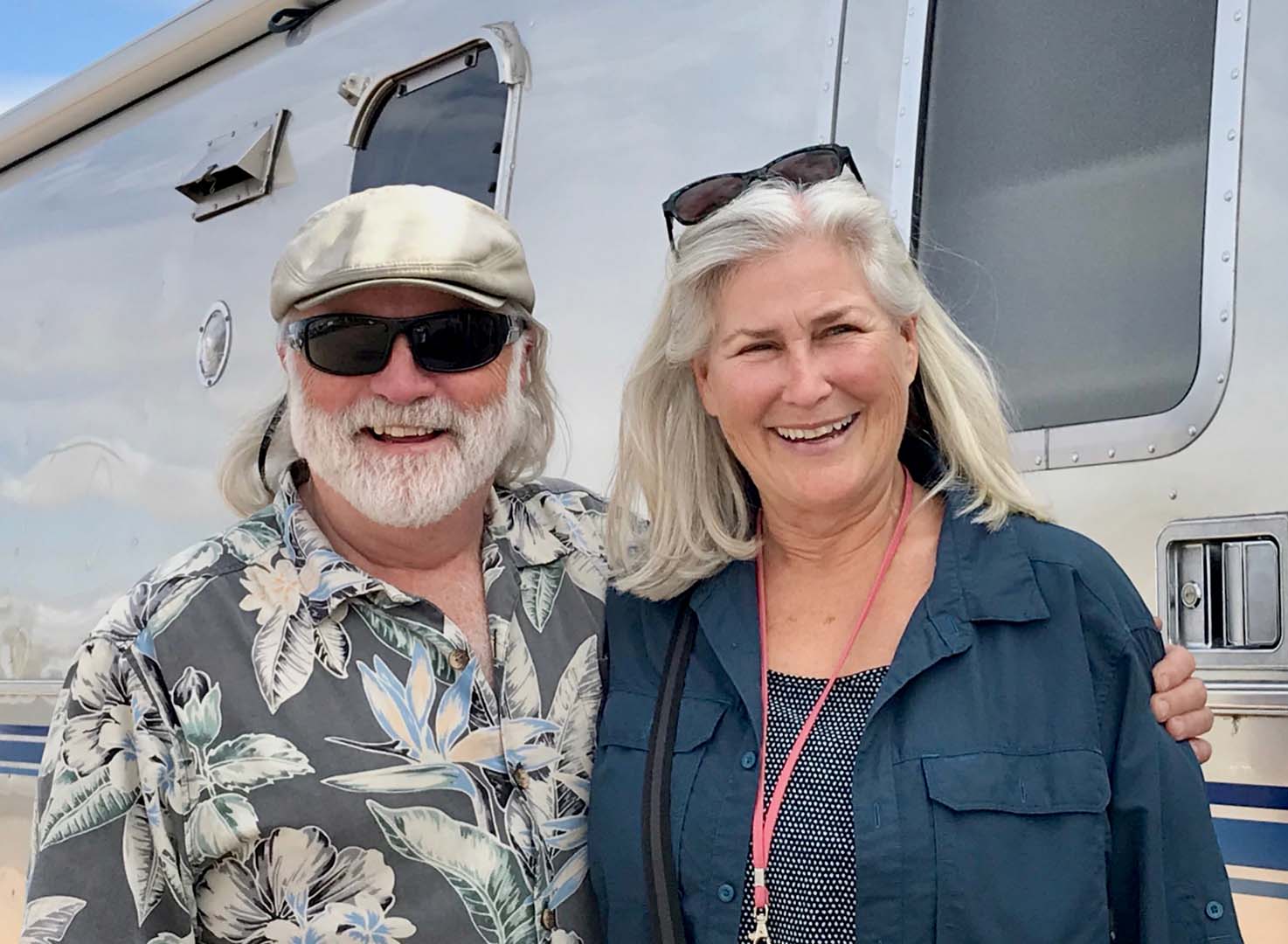 A smiling couple stands in front of a silver trailer, both wearing casual shirts, sunglasses, and hats.