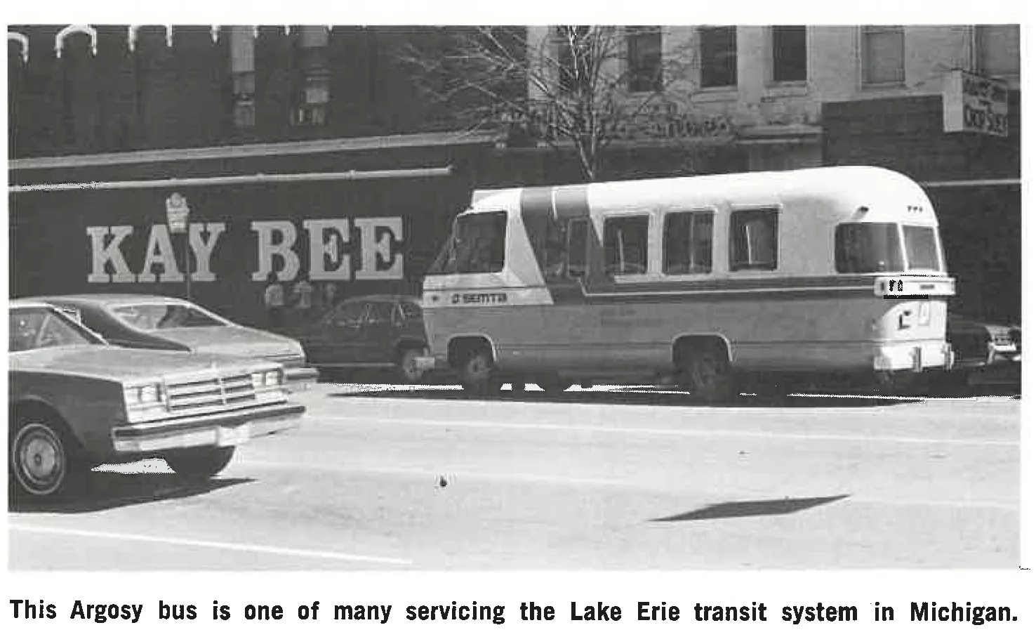 A vintage Argosy bus marked E. SMITH is parked on a street near a building with a large KAY BEE sign. A car drives past in the foreground. The scene is part of the Lake Erie transit system in Michigan.