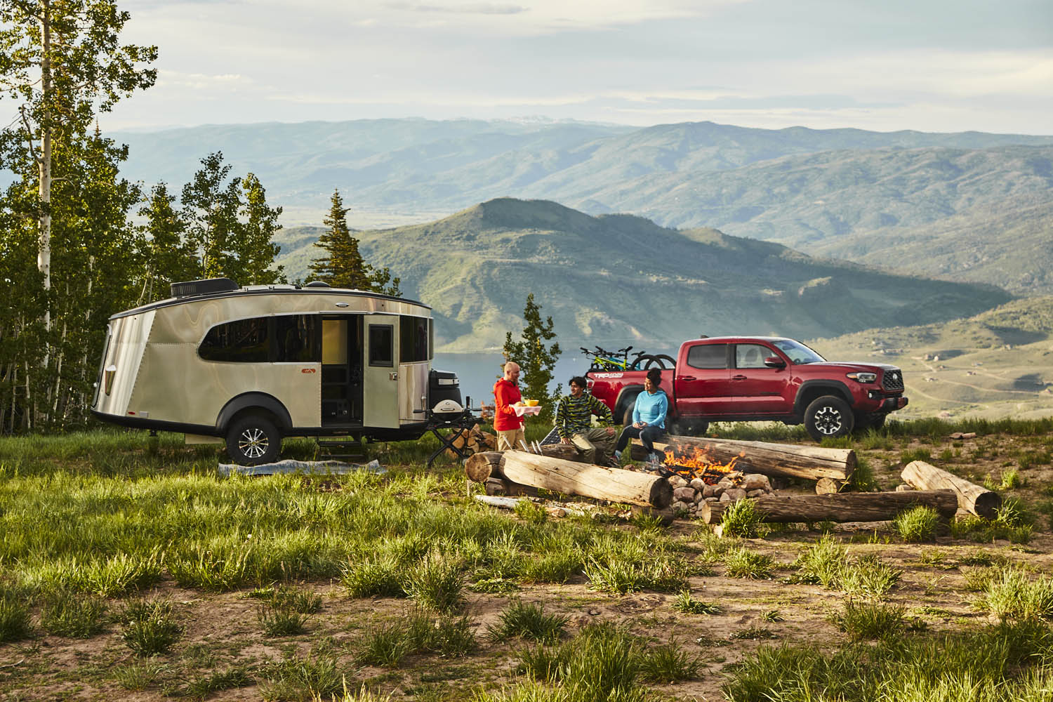 A camper trailer and red truck parked near a campfire with two people sitting, set against a mountainous landscape with trees.