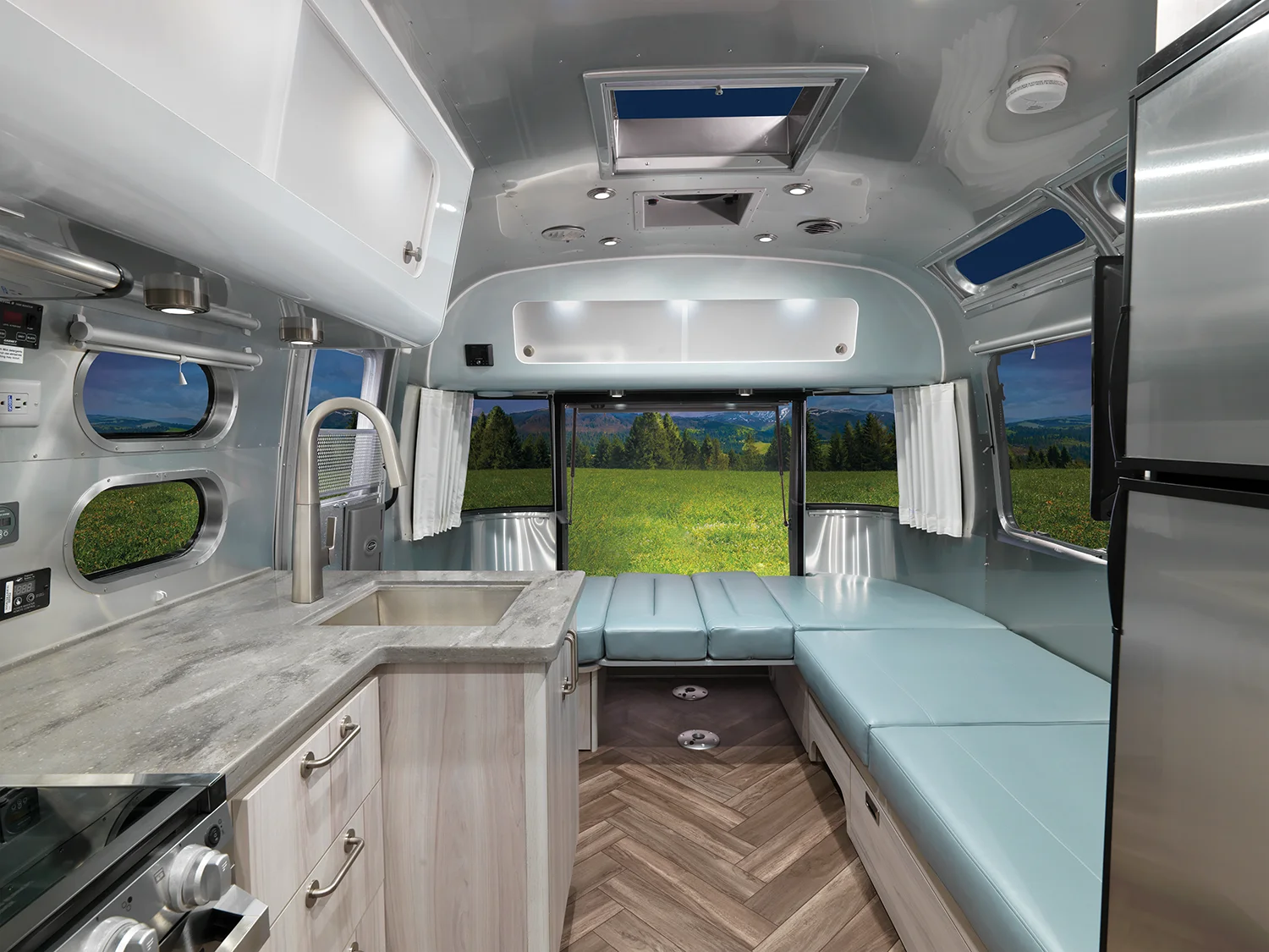 Interior of a modern silver camper van with light wood cabinets, a small kitchen, stainless steel appliances, blue cushioned seating, and large windows overlooking a green field and mountains.