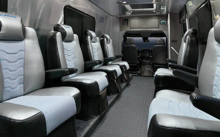 Luxury van interior with two rows of spacious, cushioned leather seats facing forward, gray and black color scheme, overhead lighting, and a driver’s area visible at the front.