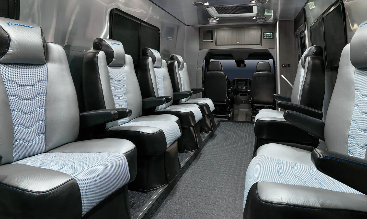 Interior of a sleek passenger van with two rows of cushioned seats facing each other, gray and black design, and a view towards the driver's area.