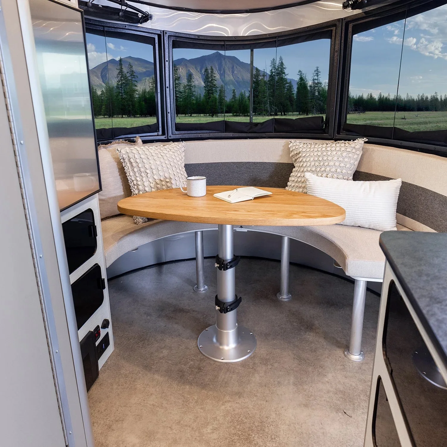 Cozy RV interior with curved seating, wooden table, pillows, and panoramic windows showcasing a scenic mountain and forest view.