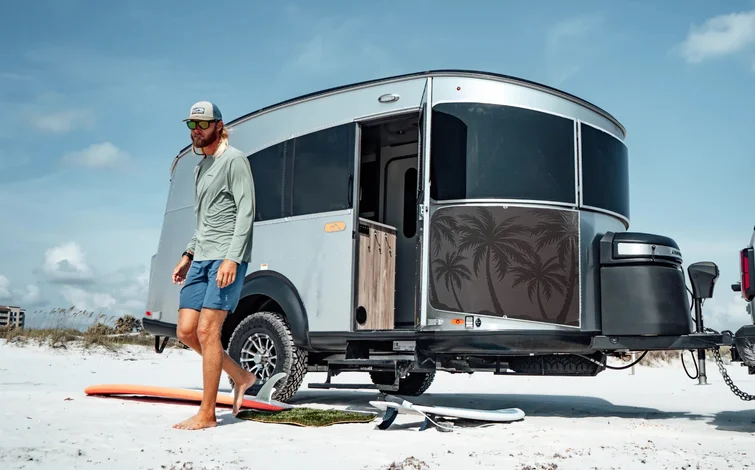 Man walks barefoot on sandy beach near a silver camper with a palm tree design, surfboards in the background under a clear blue sky.