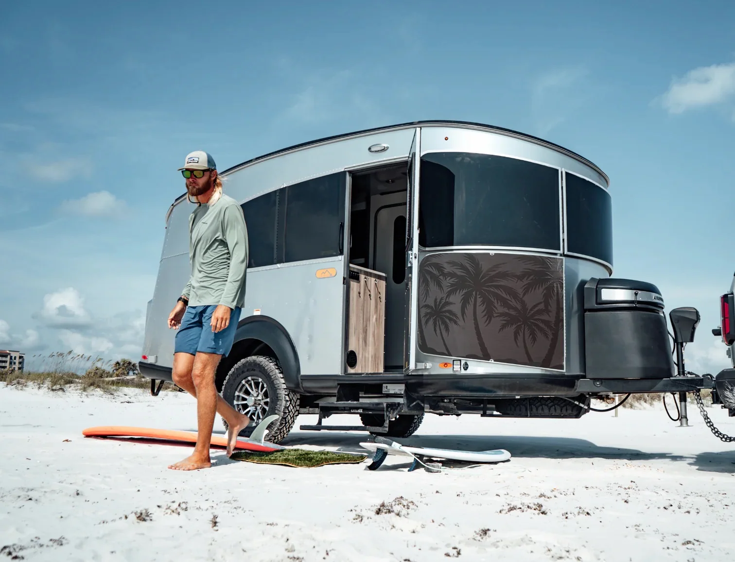 Man walks barefoot on sandy beach near a silver camper with a palm tree design, surfboards in the background under a clear blue sky.