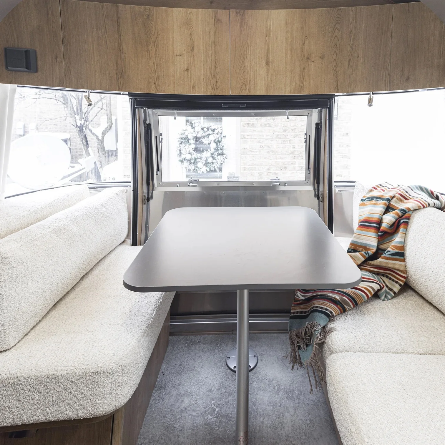 Interior of a camper with a wooden cabinet, cozy beige seating, a central table, and a colorful striped blanket near a window adorned with a wreath.