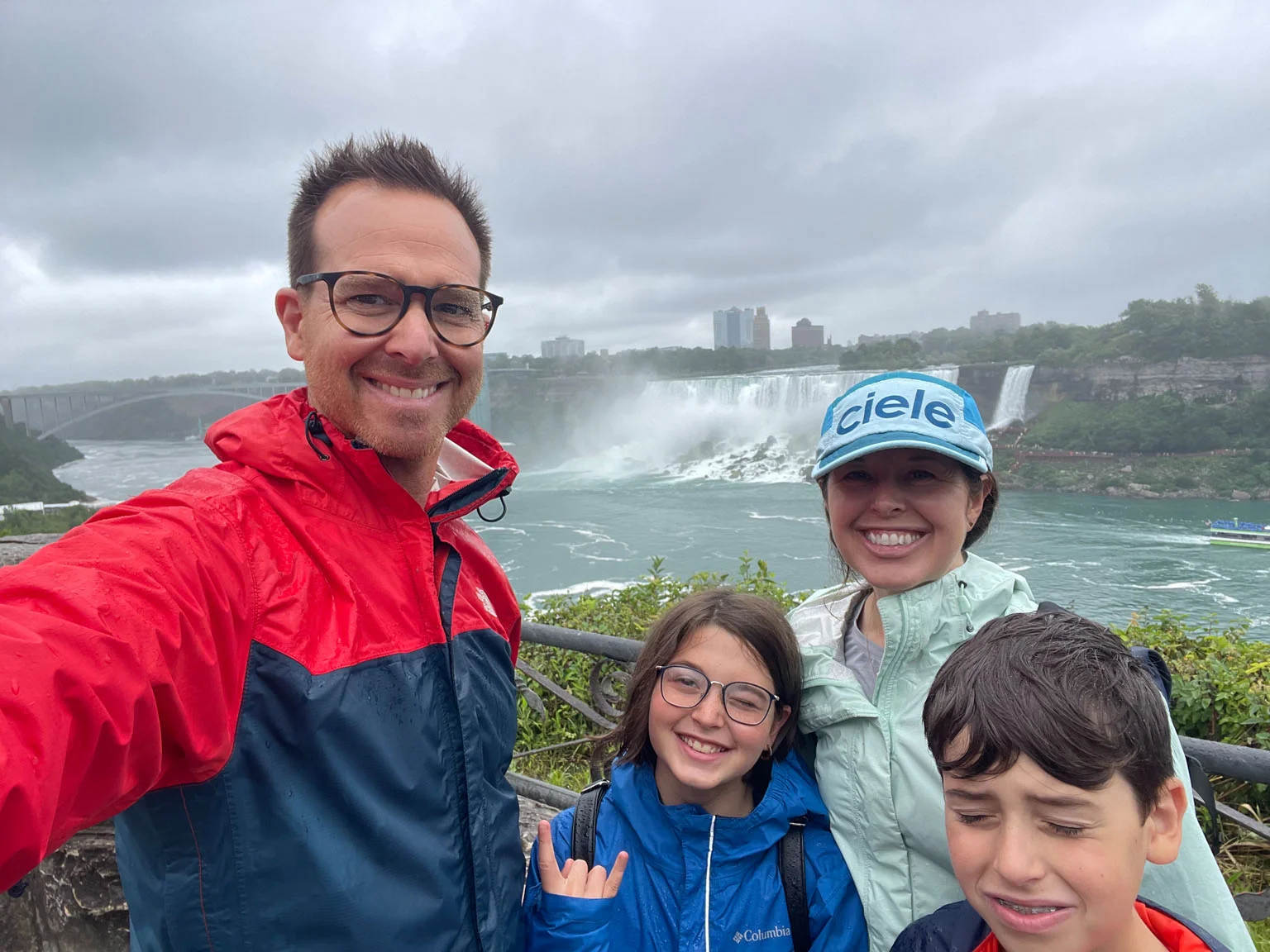 Family in rain jackets posing joyfully near Niagara Falls with misty waterfall background on a cloudy day.