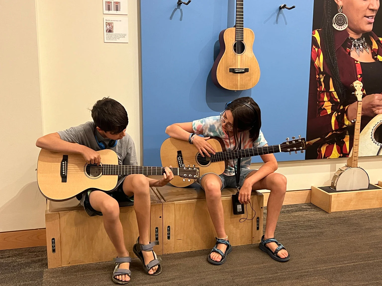Two children sitting on a wooden bench playing acoustic guitars, with banjos displayed on a blue wall behind them.