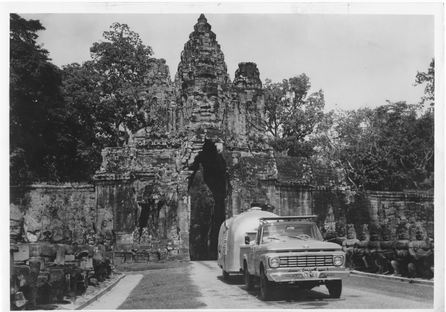 A vintage truck towing an Airstream trailer drives through the ancient stone gate of Angkor Thom in Cambodia, featuring large carved faces and flanked by statues on both sides.