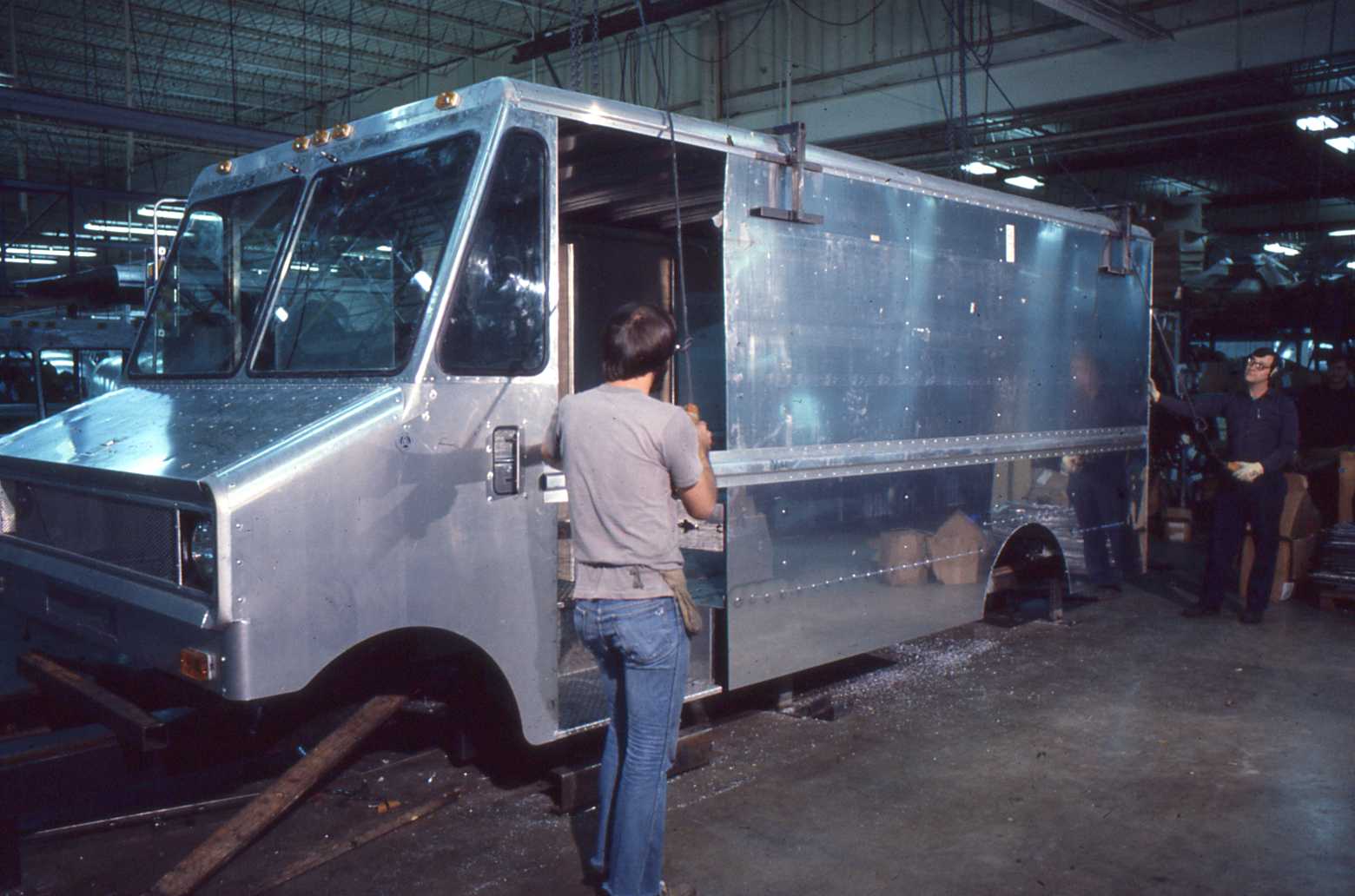 Two men work on an unpainted delivery truck inside a factory with machinery and stacks of materials visible in the background.