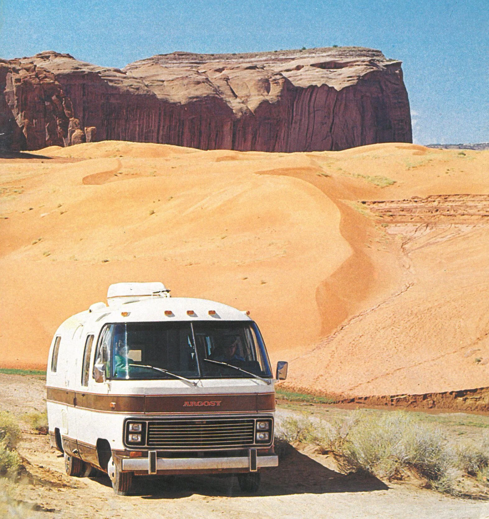 A vintage white and brown RV is parked on a dirt road in front of orange sand dunes and large red rock cliffs under a clear blue sky.
