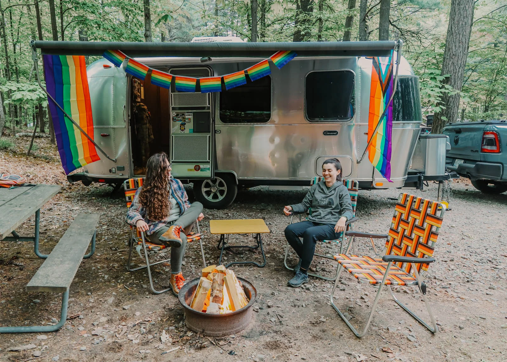 Two people sit in colorful chairs around a fire pit at a campsite, with a silver trailer decorated with rainbow flags in the background, surrounded by trees.