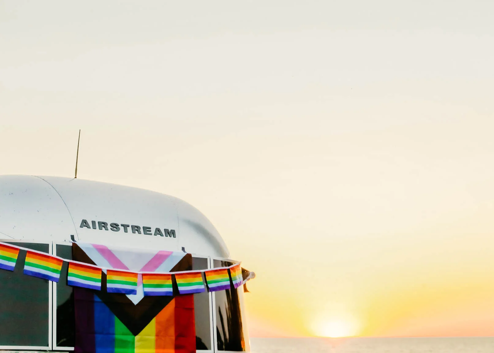An Airstream trailer decorated with rainbow and Progress Pride flags is parked by the ocean at sunset, with a bright sky and the sun low on the horizon.