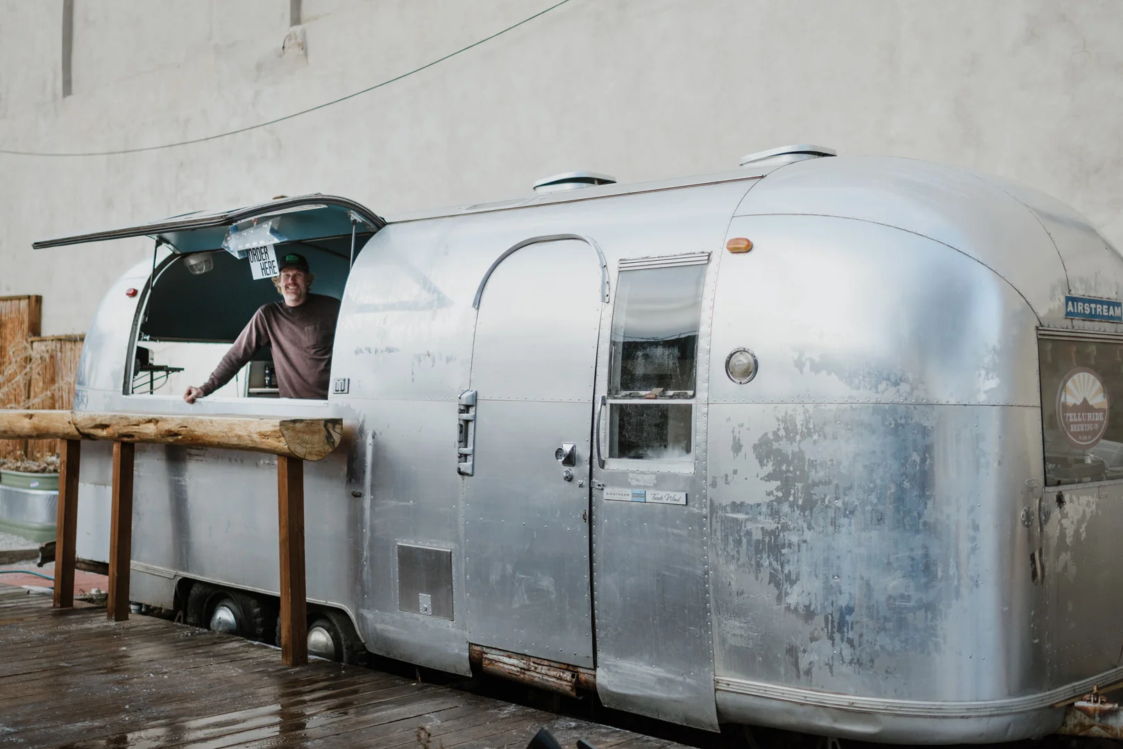 A man smiles from the serving window of a shiny silver Airstream trailer parked on a wooden deck beside a rustic wooden railing, with a beige wall in the background.