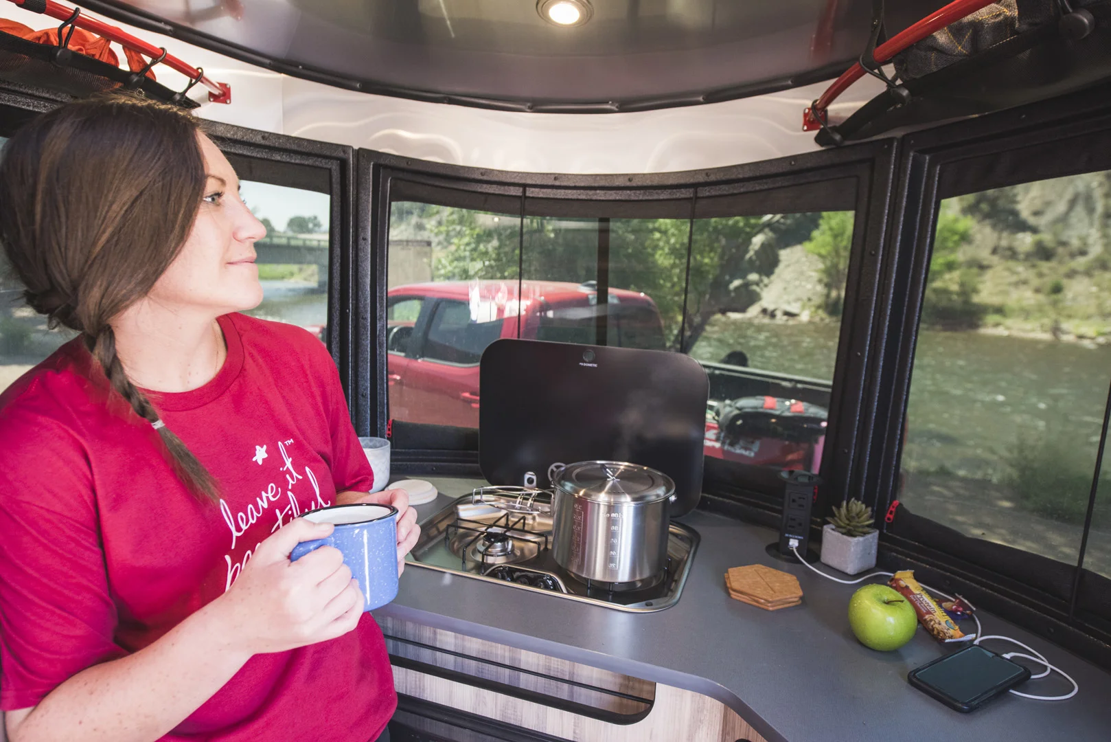 A woman in a red shirt holds a mug inside a camper van kitchen, looking outside. A pot is on the stove, and items like a phone, apple, and plant are on the counter. A river and red truck are seen through the windows.
