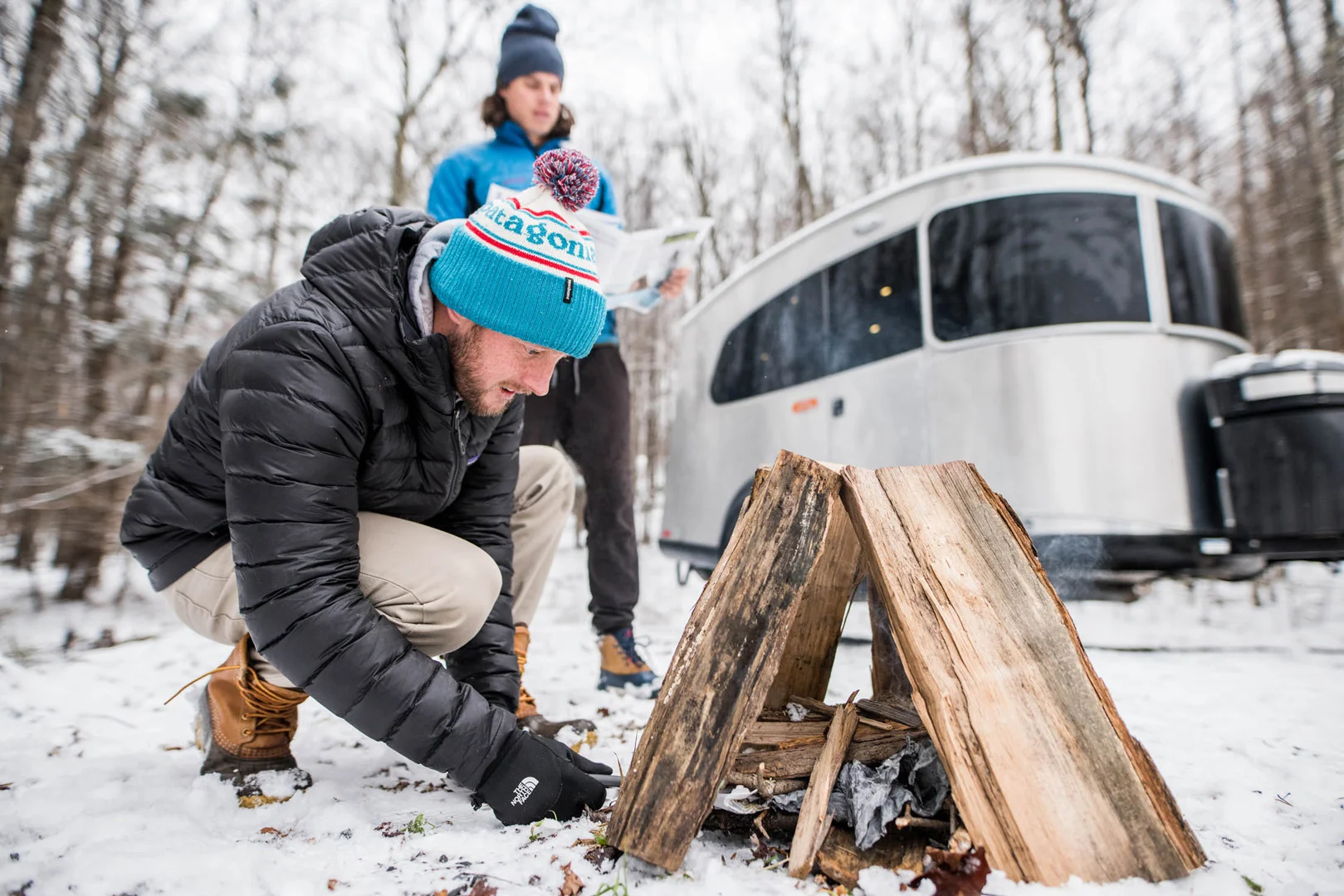Two people start a campfire in a snowy forest near a shiny Airstream trailer. One wears a black jacket and blue hat, the other holds firewood.