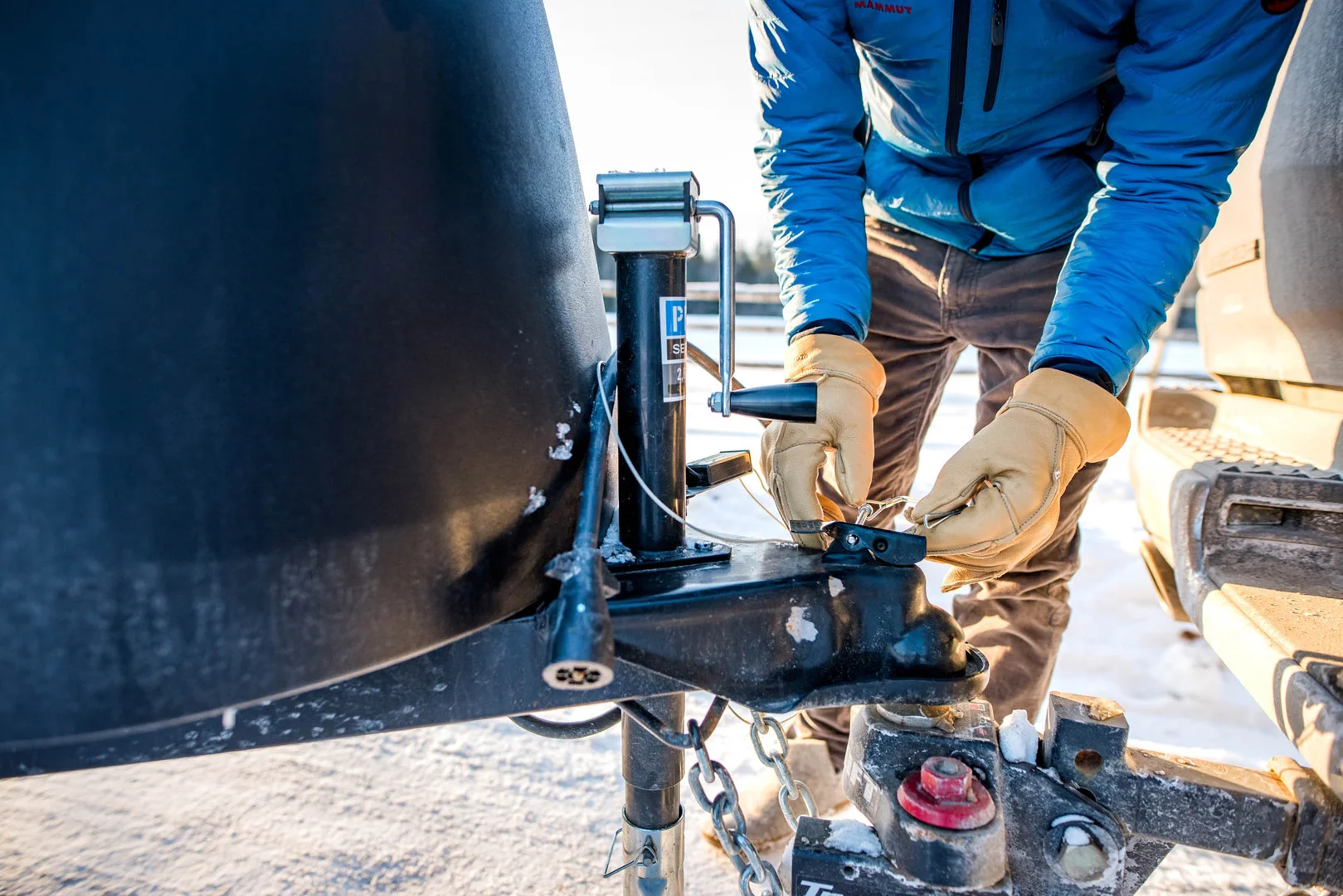 Person in a blue jacket and gloves securing a trailer hitch to a vehicle in a snowy setting.