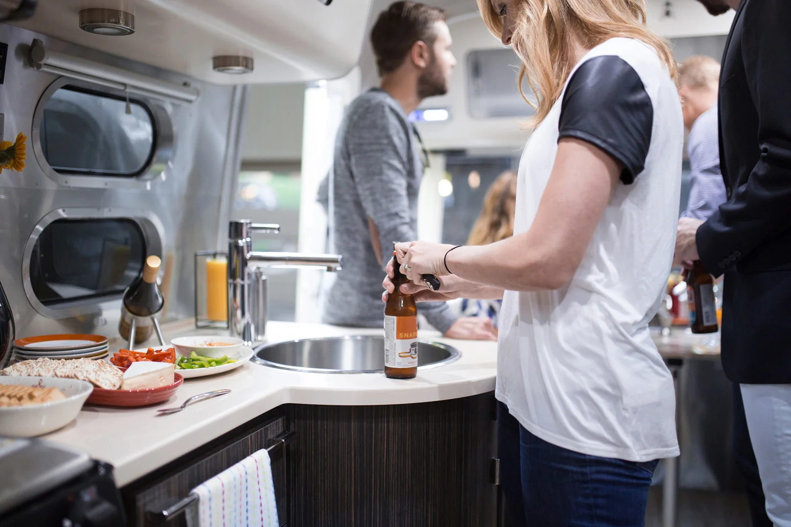 A woman opens a bottle of beer at a kitchen counter inside an RV, with food and drinks nearby. Three other people stand and talk in the background. The scene is casual and social.