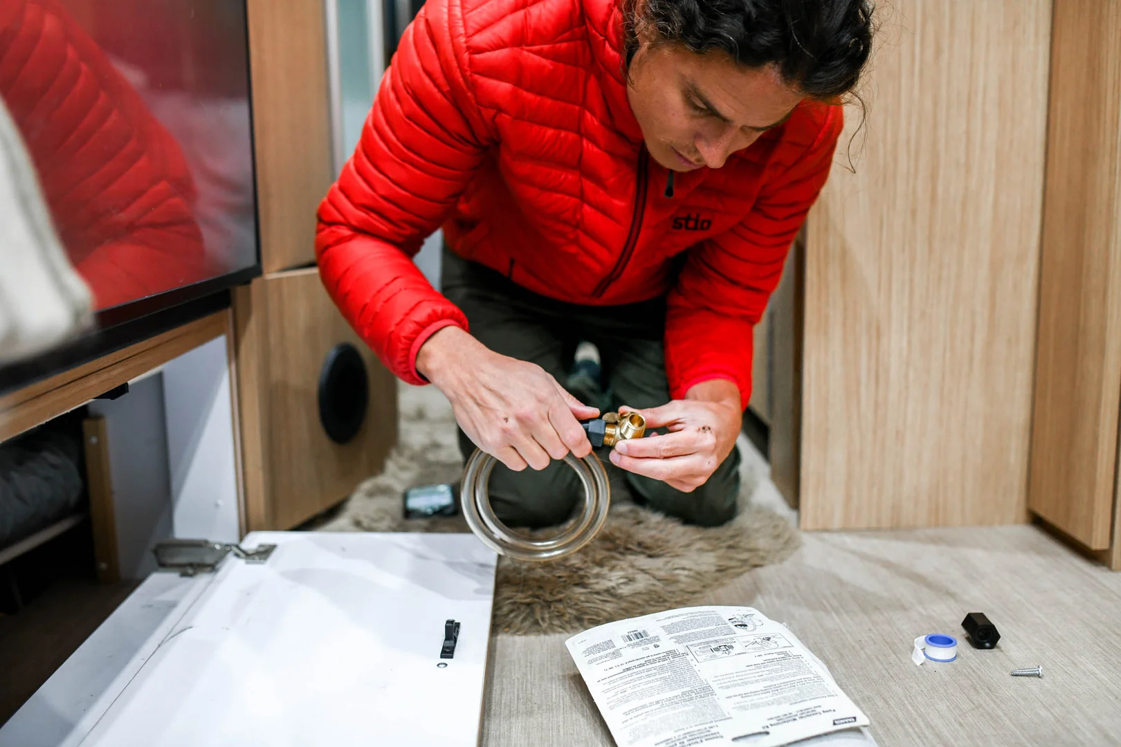 A person in a red jacket works on a plumbing project indoors, wrapping tape around a pipe. Tools, a manual, and plumbing supplies are spread out on the floor nearby.