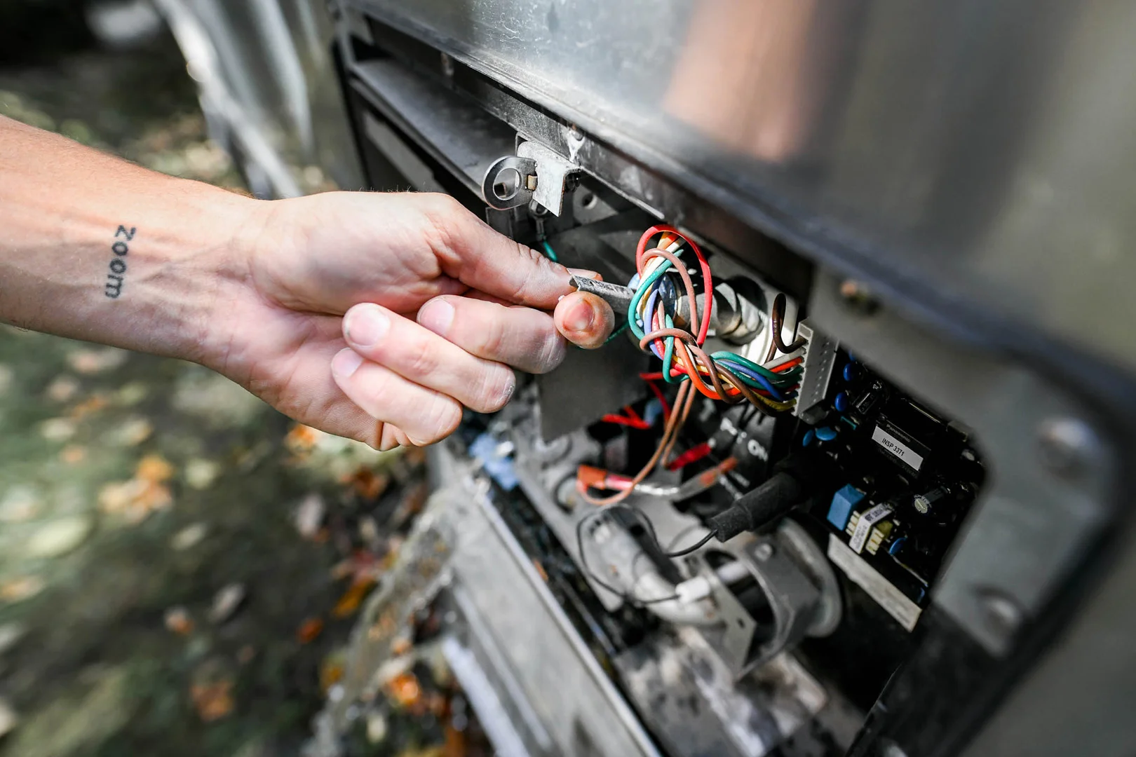 A hand holding a green wire works on a panel with colorful wires and circuits inside an open metal compartment, possibly for electrical repairs.