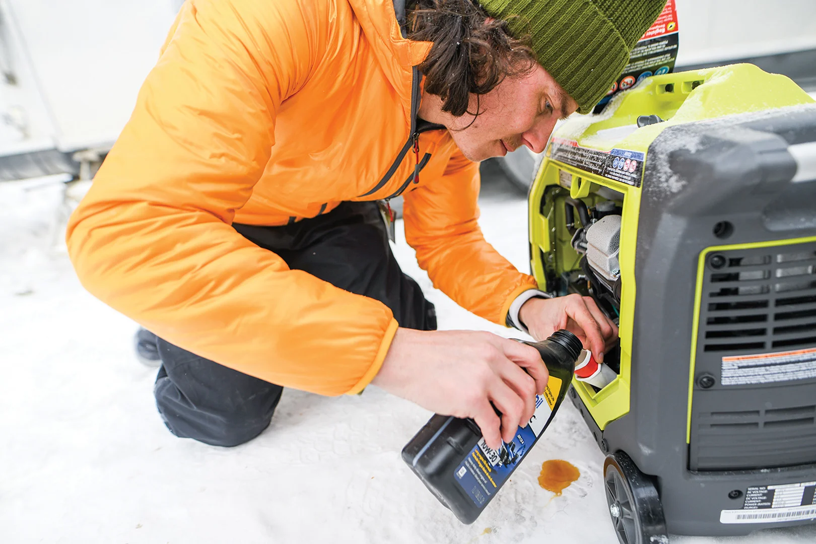 Person in orange jacket and green beanie pours oil into a portable generator on snow-covered ground.