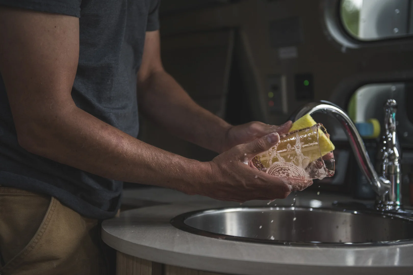 A person washes a clear glass with a yellow sponge at a kitchen sink, with water running from the faucet. The background is dimly lit, highlighting the cleaning action.
