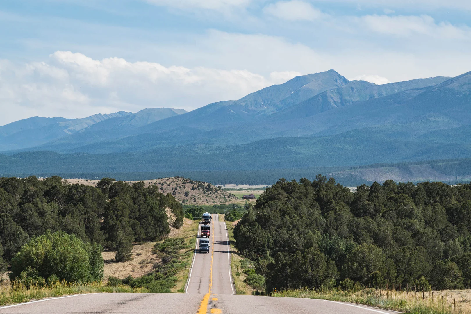 A line of cars drives down a two-lane road surrounded by trees, with mountains and a partly cloudy sky in the background.