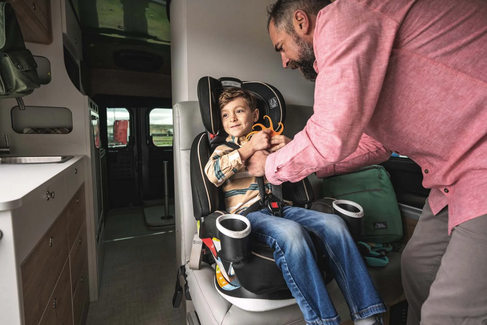 A man secures a smiling child in a car seat inside a vehicle. The child holds a toy and wears a plaid shirt and jeans.