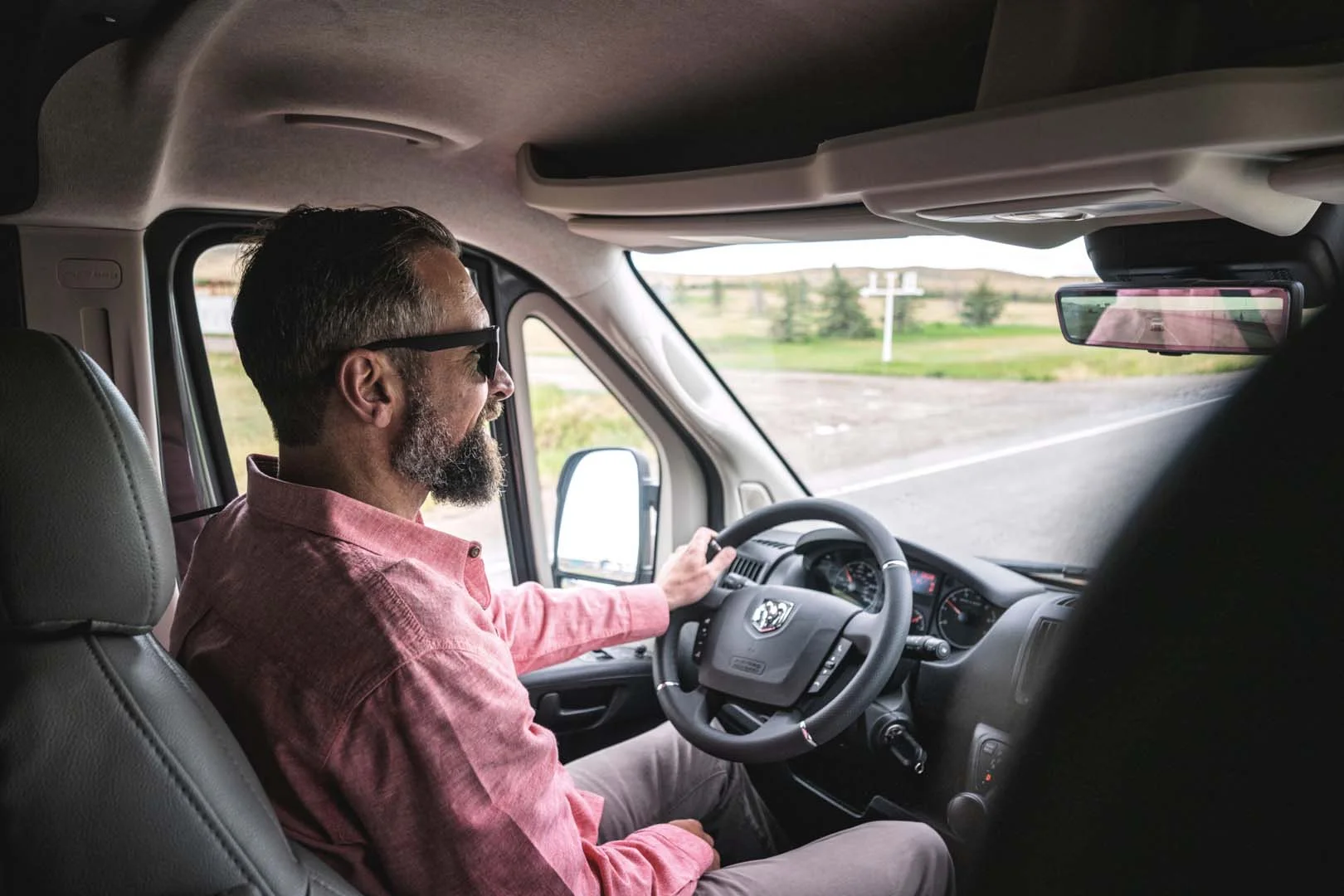 Man in a pink shirt and sunglasses driving a van on an open road, with grassy fields and trees visible through the windows.