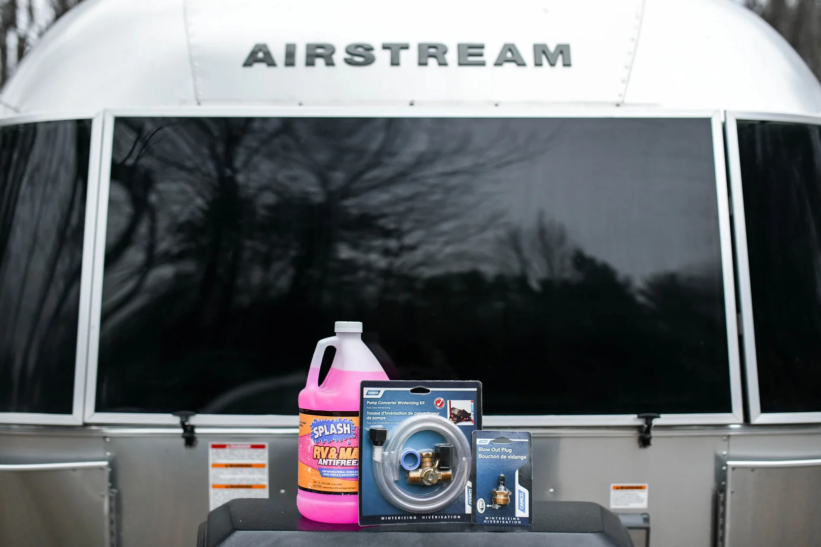 A bottle of pink RV and marine antifreeze, a brass trailer lock, and a blue pack of replacement keys are displayed in front of an Airstream trailer with a reflective window.