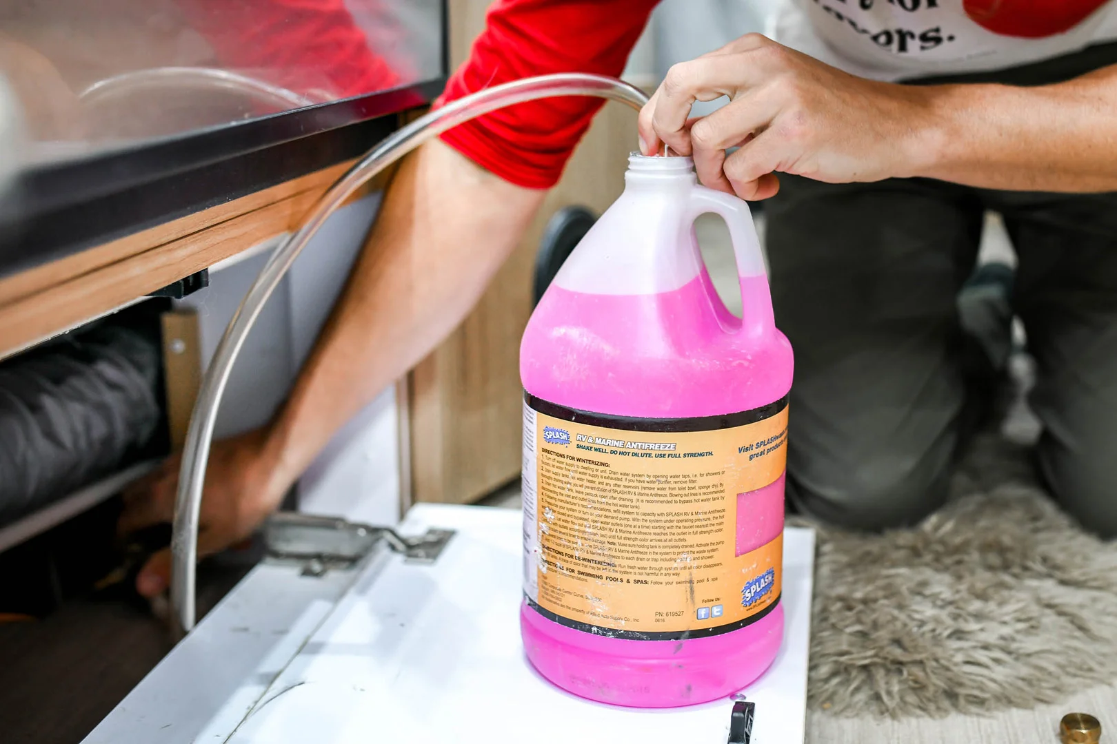 A person fills a clear tube with pink liquid from a large jug, possibly antifreeze, near an open cabinet or compartment inside a home or RV.