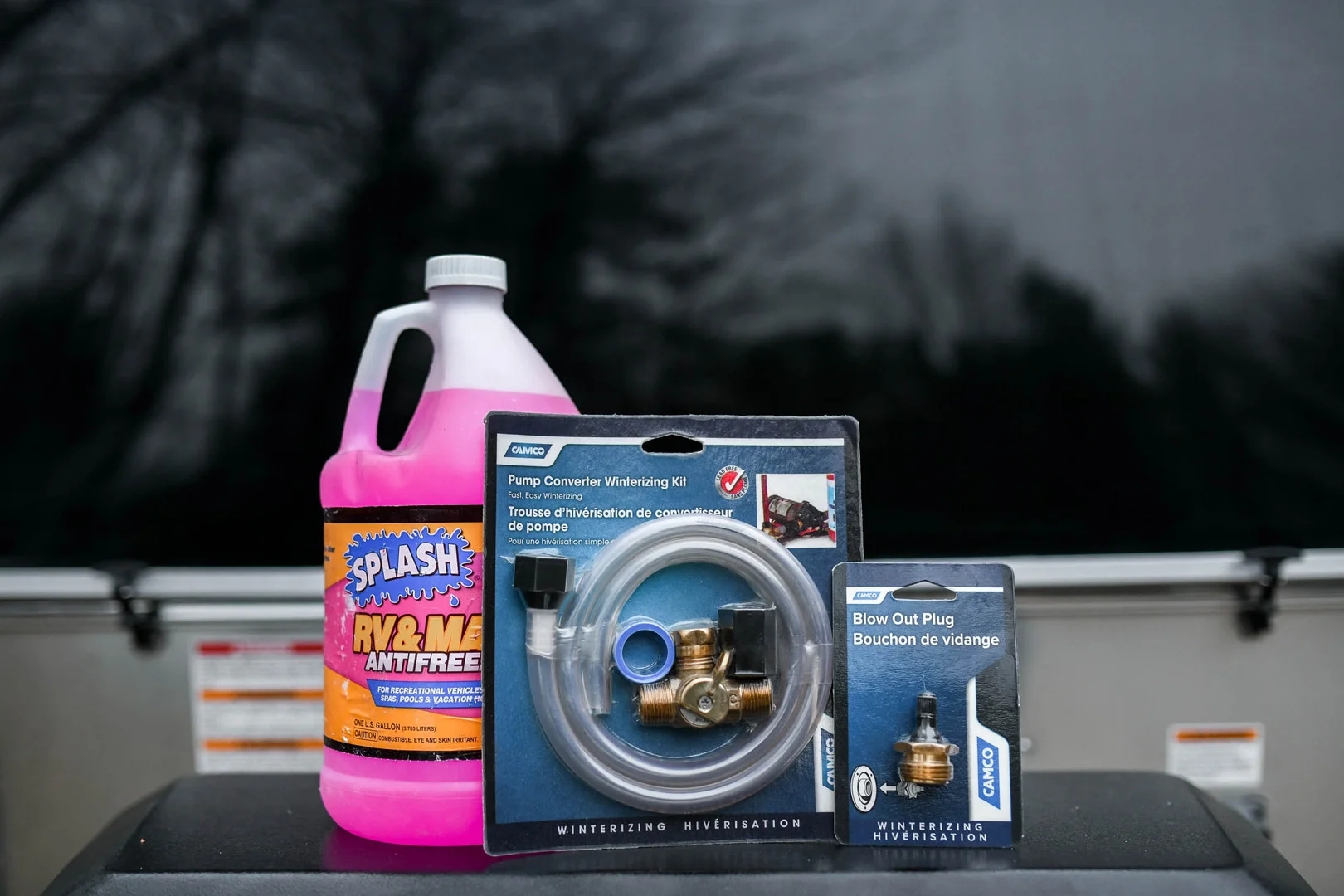 A bottle of pink RV and marine antifreeze, a pump converter winterizing kit, and a blow out plug kit are displayed outdoors on a dark surface, with a blurry background of trees and a vehicle window.